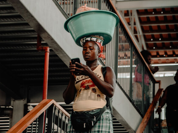 Woman in Africa reading her phone while carrying a basket on her head.